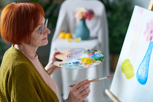 Senior red haired woman in eyeglasses looking at her picture on canvas during painting at class