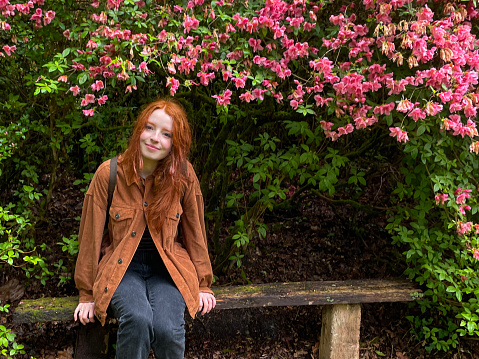 Stock photo of a young woman with long red hair sitting on a rustic style wooden bench under large rhododendron bush with pink flowers. This simple bench consists of just two posts and a thick plank of wood joining them together.