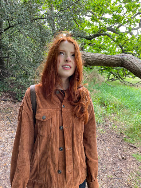 Image of attractive, red haired young woman stood looking to the sky, English Oak tree (Quercus robur) trunk on woodland floor, storm damage, focus on foreground Stock photo showing an English Oak tree (Quercus robur) that fall down several years ago, following winter storm damage. The majority of the tree's roots remained in the ground, and so the tree managed to survive and continues to grow, on its side. corduroy jacket stock pictures, royalty-free photos & images