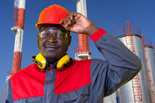Young African-American Oil Worker in Red Hardhat Standing In Front Of Oil Industry Storage Tanks.