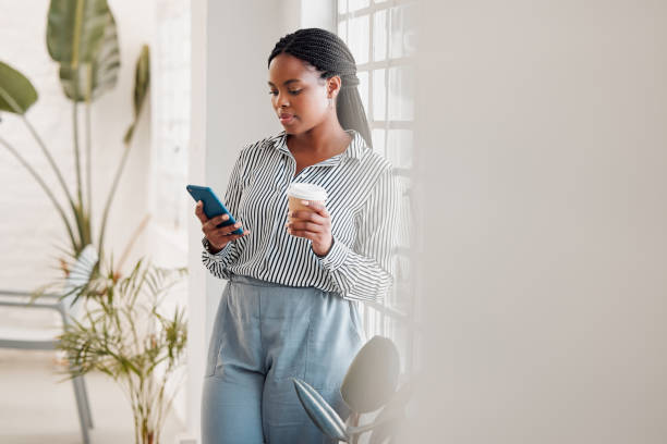 istock African american businesswoman using her phone while drinking a coffee alone in an office at work. One black female businessperson using social media on her cellphone and holding a coffee cup on a break standing at work 1399012413