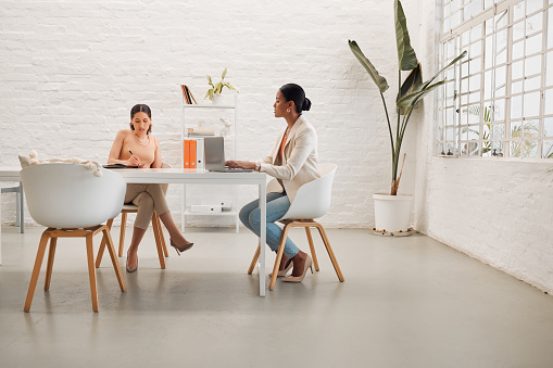 Two young mixed race businesswomen having a meeting writing notes and using a laptop in a boardroom at work. Focused businesspeople talking while planning together in an office