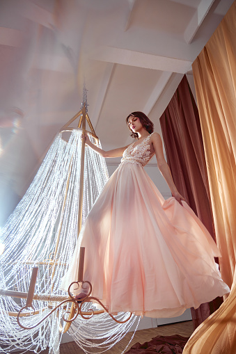 Art beauty woman in long beige pink dress dances and spins on large crystal chandelier in center of hall. Fantasy Fashion woman in a light summer dress