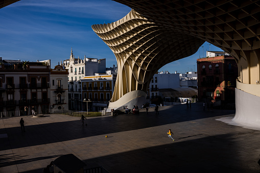 Seville, Spain, April 9, 2022: City life under the Metropol Parasol during a sunny and warm spring day on April 9, 2022 in Seville, Spain. People enjoy the shadow and freshness under the Metropol Parasol architecture. A kid runs after his soccer ball under a stripe of sun