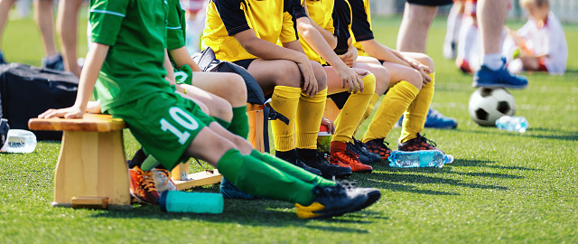 Football players are wearing soccer cleats and jersey kits in the youth team sitting on a wooden bench. Group of kids in a school sports team.