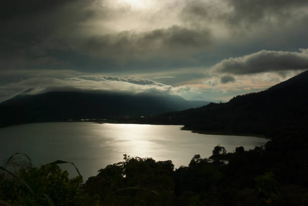 Dramatic storm in mountains landscape - heavy rain fluffy clouds and sunbeams over black mountains and bright shine lake. Tradition rain season in indonesia on Bali.