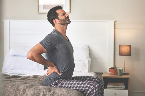 A man feeling back pain in bed at home. A mature guy looking unhappy, stressed and feeling sick in his bedroom during the morning