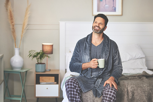 A man having coffee in bed at home. A happy mature guy thinking and sitting in his bedroom during a relaxing morning