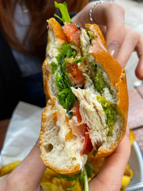 Stock photo showing close-up, elevated view of chicken pesto burger served in bread bun with chicken breast on bed of rocket leaves and beef tomato slice, topped with green pesto, served with chips on enamel plate lined with greaseproof parchment paper. imagen de primer plano de una persona irreconocible sosteniendo gourmet a medio comer, hamburguesa de pesto de pollo con papas fritas, bollo de pan con pechuga de pollo, rúcula, rebanada de tomate de res, fondo de mesa de madera, vista elevada