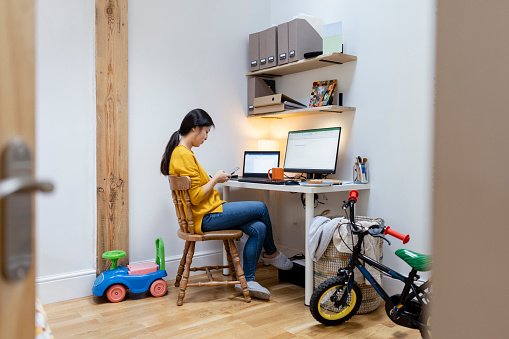 A side-view shot of a mid-adult Asian woman sitting in her home office at her desk wearing casual clothing, she is working at her computer using her smartphone. Children's toys can be seen around the room, she is a working mother.