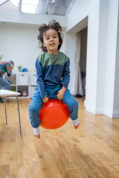 A front-view shot of a young boy wearing casual clothing bouncing on a space hopper in the living room. He is having fun with a big smile.