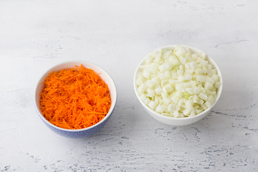 Grated carrots and diced onions in ceramic bowls on a light gray background, top view. Cooking healthy homemade food