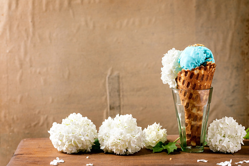 Turquoise ice cream with white flowers in waffle cone, standing in transparent glasses on wooden table. Trendy homemade summer dessert. Copy space