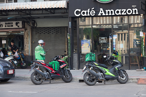 Thai express delivery person at small coffee shop in Bangkok Ladprao. Man has arrived at coffee shop branded Amazon and is on way to entrance. A second express delivery motorcycle is parked in front of shop