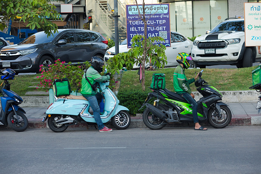 Two thai express delivery persons are waiting on their motorcycles. Men are using their mobile phones. Scene is in street Wanghin Road in Bangkok Ladprao near small shopping center complex The Jas