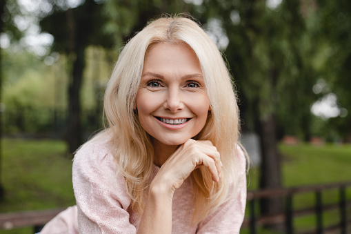 Cheerful caucasian mature blonde woman wife with toothy smile relaxing resting walking in city park forest alone looking at camera outdoors