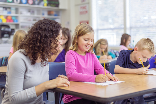 An elementary age girl smiles while receiving help from her teacher on an assignment she is working on in class.