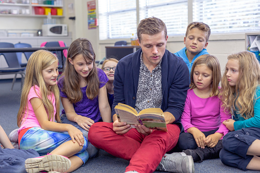A male teacher sits on the floor in his classroom and reads a storybook out loud to his elementary age students who are gathered around him listening intently.
