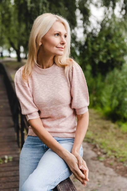 vertical portrait of a caucasian mature beautiful blonde woman wife walking alone in park forest outdoors spending free time in woods. - footpath field nature contemplation imagens e fotografias de stock