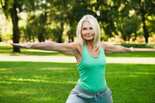 Beautiful mature fit woman in fitness outfit doing physical exercises in public park, stretching, finding body balance in yoga class.