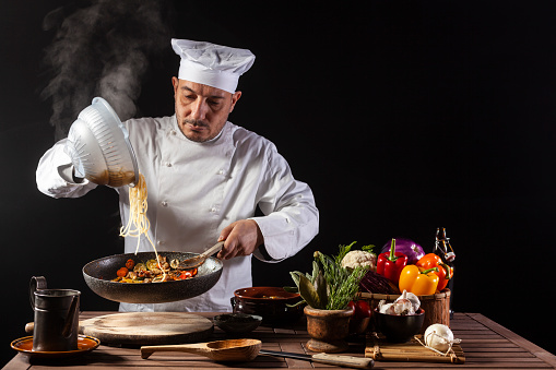 Male chef in white uniform pouring boiled spaghetti into pan wok for cooking pasta with vegetables. Backstage of preparing traditional italian dish on black background
