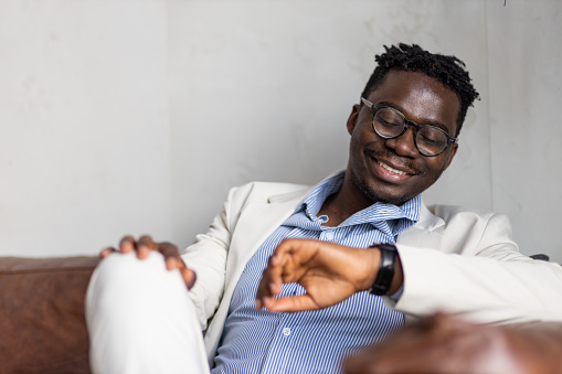 Handsome young black man in business wear sitting casually on a sofa. Young African-American businessman at work, checking the time. Portrait of a happy black man smiling, looking at his wristwatch.