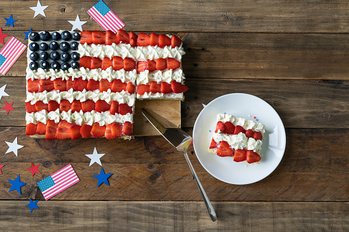 Square cake with the colors of the US flag on a wooden background with decoration. Plate with slice of cake. Independence day celebration. Top view. Copy space.