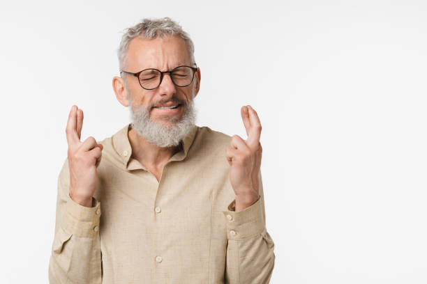 pedir un deseo. concepto de esperanza. hombre caucásico maduro de mediana edad con camisa beige y gafas cruzando los dedos con los ojos cerrados por buena suerte, orando a dios aislado en fondo blanco - fingers crossed fotografías e imágenes de stock