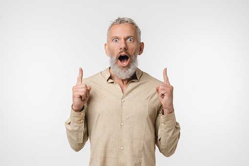 Sale discount offer. Caucasian mature middle-aged man freelancer in beige shirt pointing upwards with shock impression at copy space showing free space isolated in white background