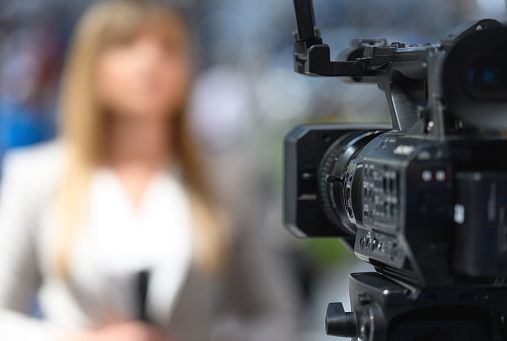 Female news reporter in front of camera, video camera is in the focus, reporter in the background is blurred, Nikon Z7