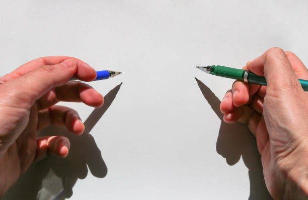 two hands of an ambidextrous young man holding a blue pen in his left hand and a green one in his right, pointing to a white sheet of paper with blank space to fill in two hands of an ambidextrous young man holding a blue pen in his left hand and a green one in his right, pointing to a white sheet of paper with blank space to fill in ambidextrous stock pictures, royalty-free photos & images