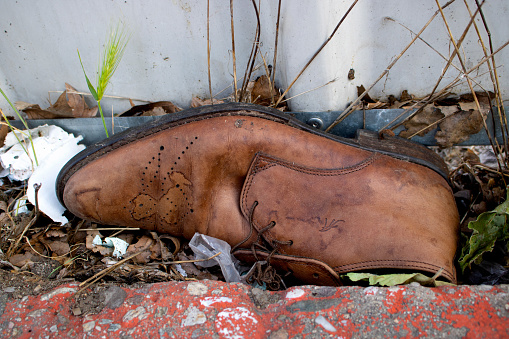 abandoned and weathered elegant brown shoe of leather among dry leaves and street garbage next to a fence - true crime scene