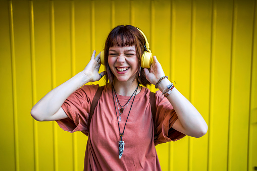 Carefree smiling young woman listening to music on headphones outdoors against yellow wall