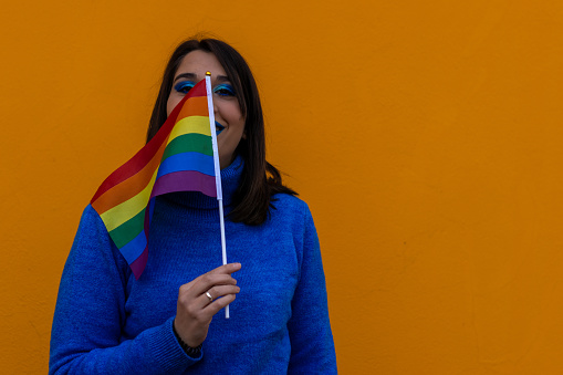 Portrait of a cheerful young woman in blue clothes on an orange background holding a rainbow-colored flag looking at camera.
