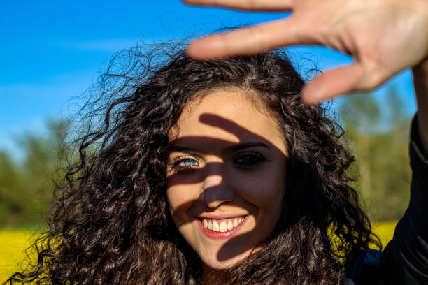 retrato de una mujer sonriente con ojos azules cubriendo el sol con la mano durante el atardecer en un campo amarillo - color de ojos fotografías e imágenes de stock