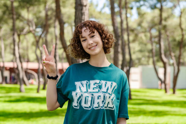 Young beautiful redhead woman wearing green tee standing on city park, outdoors smiling with happy face winking at the camera doing victory sign or gesture, showing peace. Number two. stock photo