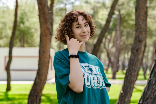 Beautiful redhead woman wearing green tee standing on city park, outdoors smiling doing phone gesture with hand and fingers like talking on the telephone. Communicating concepts. stock photo