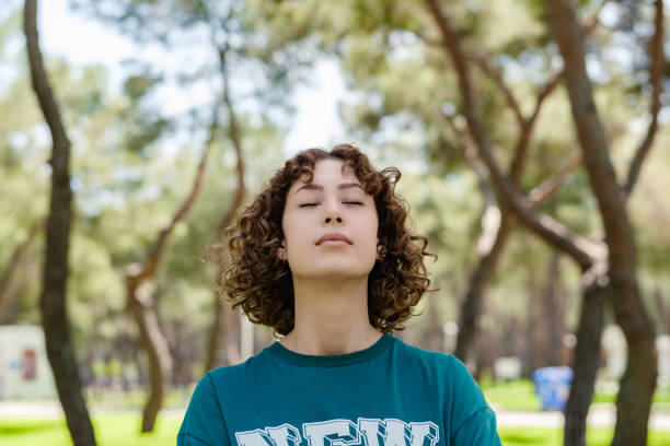 Young redhead woman wearing green tee standing on green city park, outdoors. Enjoying stress free mindful moment, doing yoga relaxation exercises, closed eyes, meditating near nature, taking breath. stock photo
