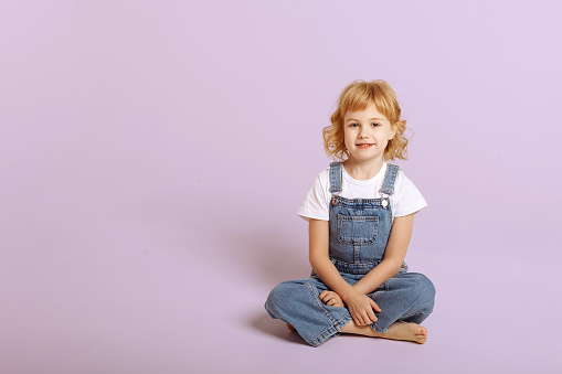 Pretty long-haired girl in a denim overalls sits cross-legged on a light pink studio background. Looking at the camera, front view, copyspace.