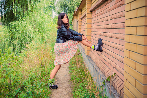 A beautiful young girl in a skirt and a black jacket stands, put her foot on a brick wall