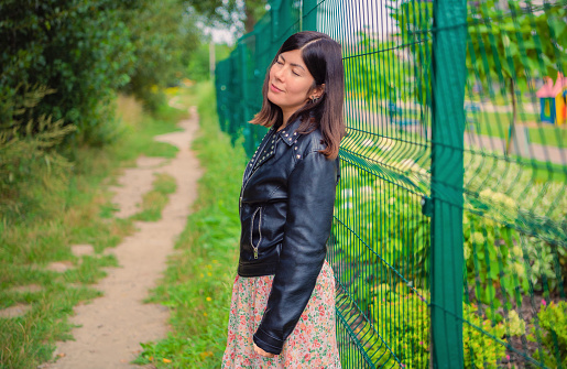 A cute girl in a black jacket stands with her eyes closed, leaning her back against a green iron fence.