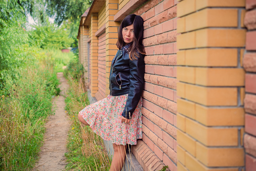 A brunette girl stands leaning back against a brick wall in a green forest