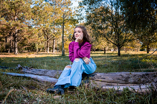 Green-eyed girl wearing blue jeans and black boots with a pink blouse sitting on a tree trunk in the middle of nature. Mention of young and beautiful women in the countryside and outdoors.