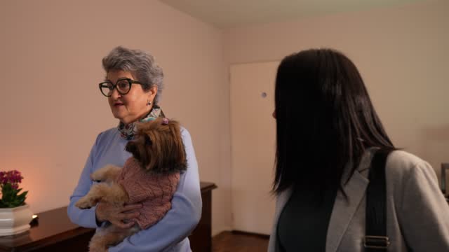 Elder woman holding a dog welcoming a woman in her house