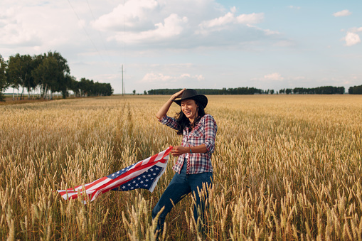 Woman farmer wearing cowboy hat, plaid shirt and jeans with american flag at wheat field.