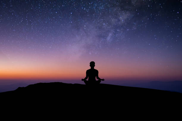 Silhouette of young female sitting practices yoga and meditating in lotus position alone on top of the mountain with night sky, star, Milky Way. She felt calm and happy. stock photo