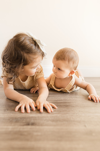 A 3-Year-Old Toddler Girl and Her 5-Month-Old Baby Brother Looking at Each Other While Laying on Their Bellies on the Floor for Tummy Time While Wearing Matching Yellow Plaid Outfits.