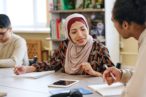 Muslim woman wearing hijab sitting at table in library taking look into notebook of her classmate during lesson for immigrants