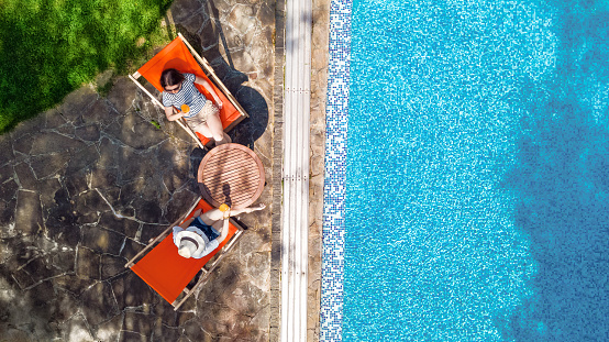 Young girls relax near swimming pool in sunbed deckchairs, women friends relax on tropical vacation in hotel resort, aerial drone view from above