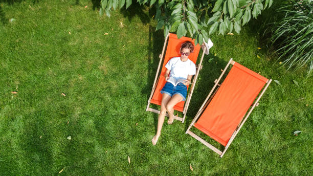 jovem relaxa no jardim de verão em deckchair sunbed na grama, mulher lendo livro ao ar livre no parque verde no fim de semana, vista aérea drone de cima - espreguiçadeira - fotografias e filmes do acervo